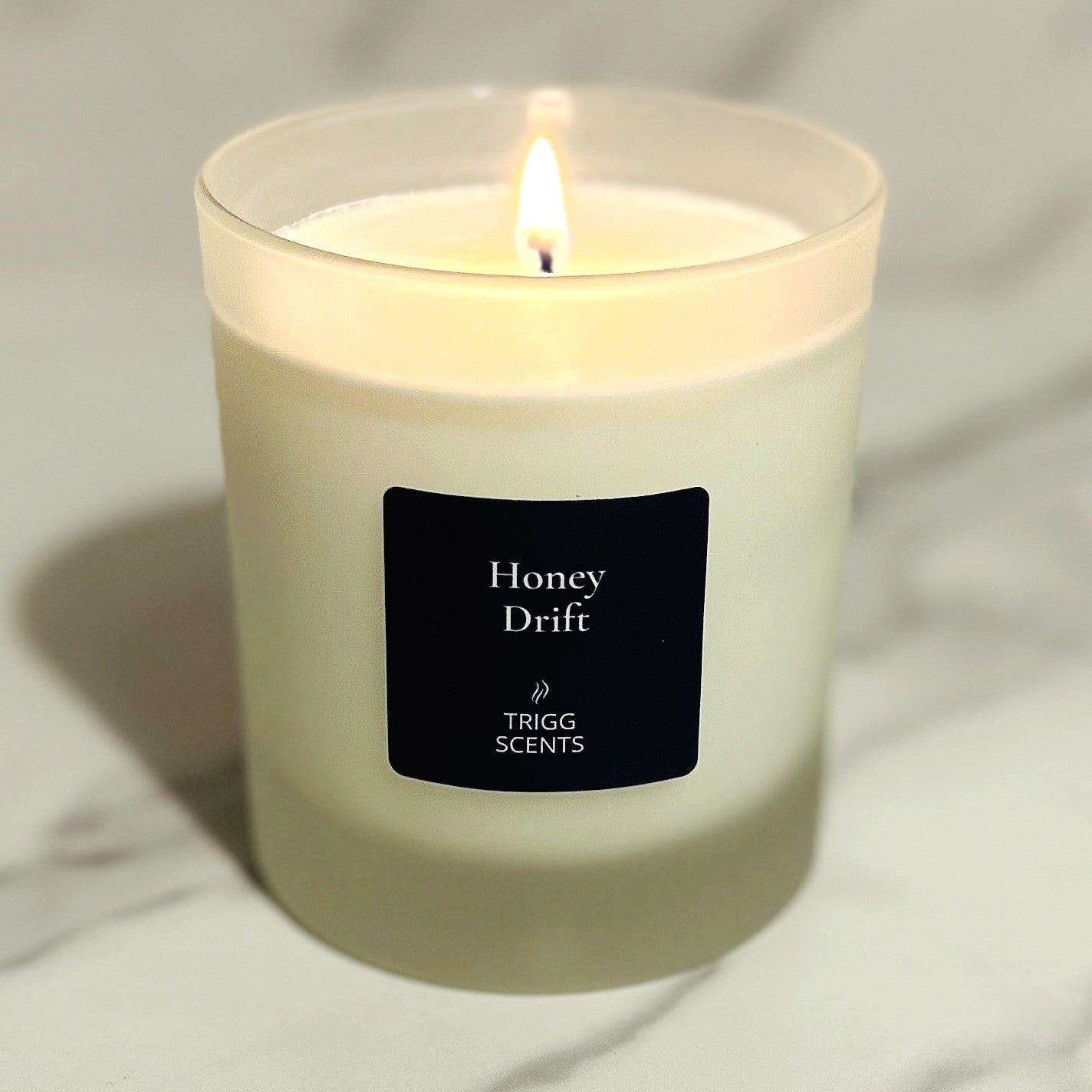 Lit Honey Drift scented candle in frosted glass jar, warm glow with black label, photographed on a light neutral background for a cosy evening atmosphere.